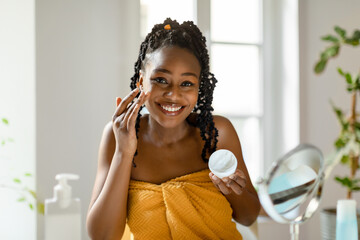 Pure joy of skin hydration. Smiling black woman applying moisturizing face cream, sitting in front of mirror in bedroom