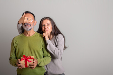 Glad caucasian elderly lady closes eyes to husband, make shh sign, gives gift, congratulations on...
