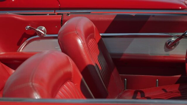 Red leather interior of old American coupe convertible car with chrome details, chairs without headrests. Manual window handles gleam in sunlight, old leather on backs has been rubbed to shine.