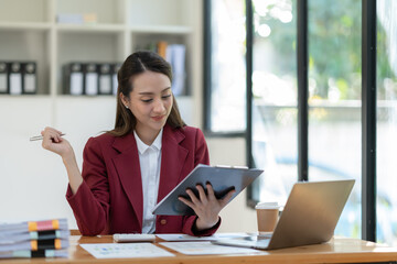 Beautiful attractive Asian businesswoman holding a clipboard of documents sitting analytically analyzing various marketing management information the finances were at her desk in the office at ease. © crizzystudio