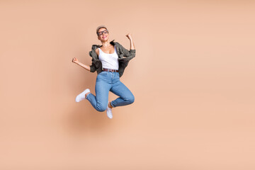 Full length photo of positive lucky lady dressed khaki shirt jumping high rising fists emtpy space isolated beige color background