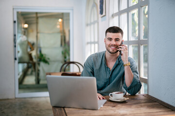 A male caucasian, Freelance bearded man on phone with laptop sitting at table smiling and looking...