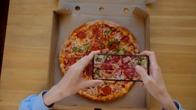 Top view - Food blogger and blogging concept. Woman in blue sweatshirt use smartphone, take photo for social networks appetizing fresh cooked italian pizza with sausage in delivery box on wooden table