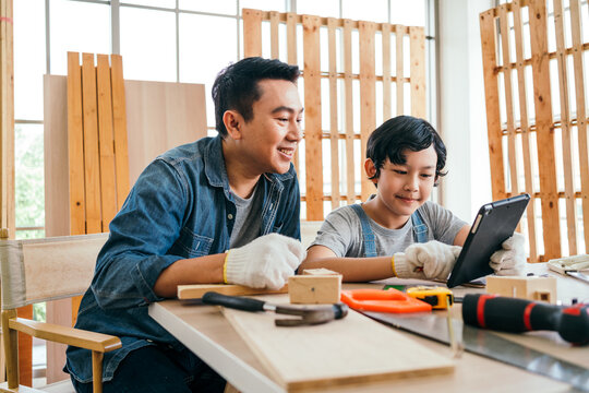 Happy Asian Father And Son Work As A Woodworker And Carpenter, Watching Content On A Digital Tablet For Making A New Woodwork Project Together. Carpentry Working At A Home Workshop Studio.