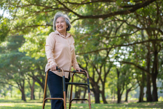 Happy old elderly Asian woman uses a walker for osteoarthritis rehabilitation physiotherapy in park.  Concept of happy retirement With care from a caregiver and Savings and senior health insurance