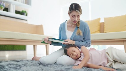 Asian mother reading a book and looking little daughter sleep on her lap in living room at home....