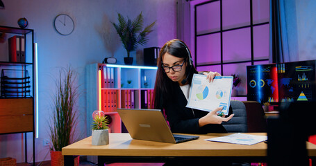 Attractive confident purposeful skilled young brunette in headset sitting in front of computer during video meeting with colleagues and explaining report with charts