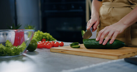 Unrecognizable woman standing near kitchen table and cutting fresh cucumber on cutting board while preparing vegetable salad