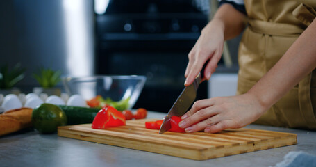 Unknown housewife slicing on small parts red pepper on cutting board using knife while preparing fresh vegetable salad for her family on dinner
