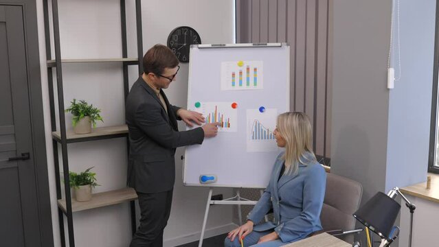 Confident business coach gives presentation on flip chart. A young woman and a man are talking and discussing business project schedules in the office