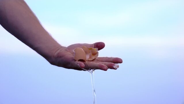 Person hand crushes brown chicken egg against blue sky