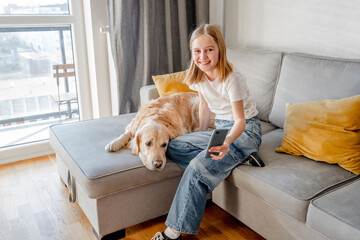 Preteen girl hugging golden retriever dog and holding smartphone on sofa at home. Beautiful child kid with labrador doggy pet using cell phone for social media connection and chatting