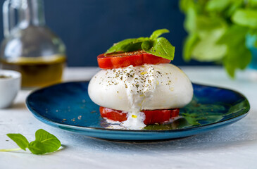 Burrata buffalo cheese with tomatoes, fresh basil, olive oil and dry oregano on a blue plate over white wooden table with ingredients and dark blue background. Close up