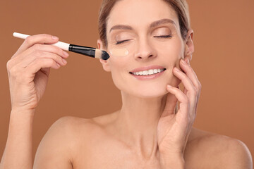 Woman applying foundation on face with brush against brown background