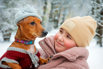 In winter, in a snowy forest, on a rainy day, a girl holds a taksus dog in her arms in a hat and clothes. The girl tenderly looks at the dog.
