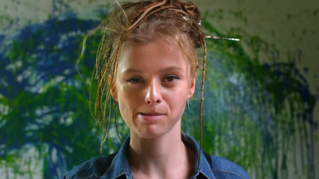 Portrait: A young talented artist with dreadlocks and red hair in an art studio. Looking into the camera, a modern painter creating abstract art. The concept of lifestyle, hobby and occupation