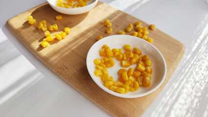 Lot of pieces of canned yellow corn on plate which is on wooden bamboo cutting board on white background. The concept of cooking and delicious healthy food