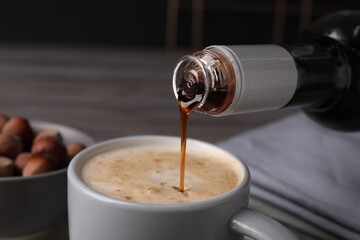 Pouring delicious hazelnut syrup into cup with coffee at table, closeup - obrazy, fototapety, plakaty