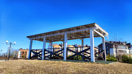 Fototapeta premium Beautiful Wooden gazebo outdoor on a Sunny autumn or spring day with blue sky. Early sprint time and pavilion or alcove in village or small town on nature landscape