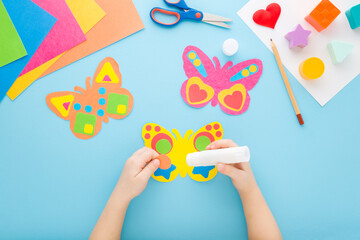 Little child hands holding glue stick and gluing colorful butterfly shape from paper on blue table...