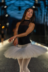 slim and young Hispanic ballerina in classical ballet costume with white tutu and black bodysuit, posing artistically with her arms crossed at night on a bridge illuminated by lights on the floor.