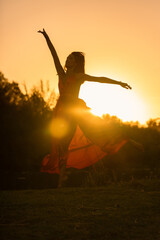 young woman in a red dress jumping in the forest at sunset against the light with the sun glinting...