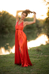 beautiful latin woman holding her hair, wearing an elegant red dress, in a beautiful natural landscape by a lake at sunset with the sunlight reflecting on the water.