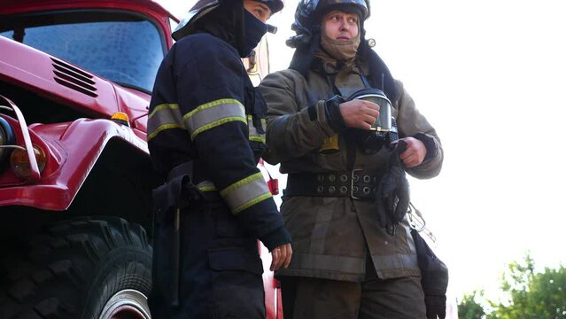 Firefighter chief talking with his subordinate about work against background of a fire engine. Young professional firemen discussing the situation report after emergency near a big red truck. Slow mo