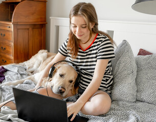 Pretty girl with golden retriever dog, headphones and laptop in bed. Female teenager with purebred doggy pet labrador making video call at pc online - Powered by Adobe