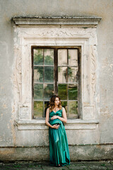 Fototapeta na wymiar Pregnant woman wearing long green dress, holding tummy near old window. New life concept.