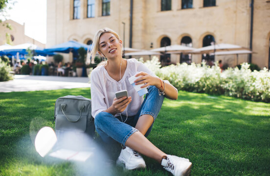 Portrait of pretty Caucasian woman in headphones looking at camera while listening audio podcast in summer park, carefree millennial female blogger with smartphone device enjoying music playlist