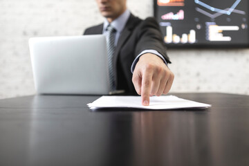 Close-up photo of male hand pointing index finger on documents. Businessman in black suit sitting at the desk and working on laptop in modern office