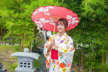 Young woman wearing traditional Japanese kimono or yukata holding an umbrella in a park with bamboo...