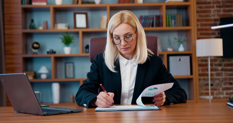 Beautiful focused experienced blond businesswoman working in office room with important financial information using computer and records in documents