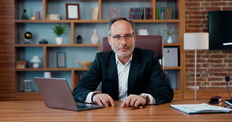 Portrait of attractive confident experienced purposeful bearded ceo in business suit which posing on camera during his work on laptop in modern office