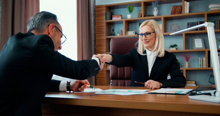 Attractive positive experienced adult businesspeople consulting about paper contract and shaking hands after reaching agreement in office