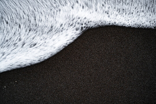 Gischt der Wellen am Strand im dunklen Sand als entsättigtes Hintergrundbild 