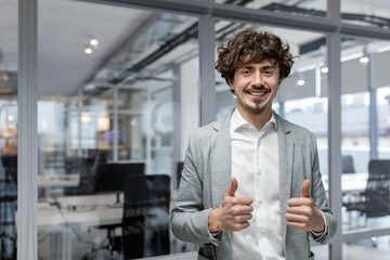 Fototapeta premium Portrait of successful businessman boss, manager in business suit and beard looking at camera and smiling standing near window, showing thumbs up, sign of success and achieving goals