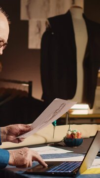 Vertical video Senior couturier work on items design with sketches, using laptop to review material and custom made sizes. Male professional tailor creating luxury clothes in workshop atelier.
