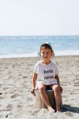 Fototapeta na wymiar Cute little toddler wearing t-shirt with lettering Hola Mom is sitting on a beach during sunny summer day