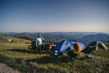 A pair of motorcyclists, man drink tea metal cup. Loving couple of tourists. Adventure, active lifestyle concept. A touring motorcycle, tents. Folding chairs. Top mountains. Ukraine