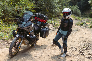 Fototapeta na wymiar Biker girl in special motorcycle outfit, protection, knee pads and turtleneck. White helmet. Stands next to a long-distance touring motorcycle. Side bags and luggage. mountain road