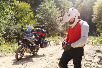 Male biker, motorcyclist in a white helmet. Stands next to a long-distance touring motorcycle. Side bags and luggage. mountain road Sunny warm summer day. Active extreme recreation.