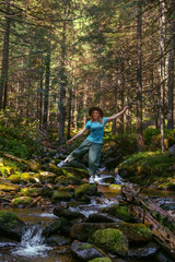 Naklejka na ściany i meble A happy woman yoga, magical, and fantastic places around the world, surrounded by nature. Female hiker crossing the river. Coniferous forest, tall Carpathian spruces. Vertical photo
