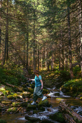 A woman explores new, magical, and fantastic places around the world, surrounded by nature. Female hiker crossing the river. Coniferous forest, tall Carpathian spruces. Vertical photo
