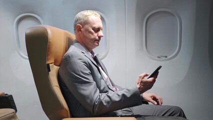 Middle aged caucasian businessman in suit sitting on seat in airplane using smartphone while business and traveling flight