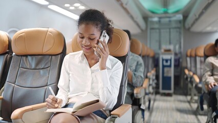 Young african american woman sitting on seat in airplane and talking by smartphone and writing on book. Young woman traveller talking on cell phone