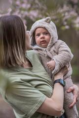 Fototapeta premium A charming boy in nature among flowering trees in his mother's arms. A child of 4 months plays with his mother and smiles. Family, pregnancy and childbirth.
