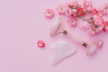 Facial massage kit for home spa. Face roller and gua sha massager made from rose quartz on pink pastel background with rose buds. Natural treatment concept.