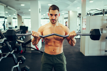 Portrait of a young muscular man lifting a barbell in a gym while looking at the camera.